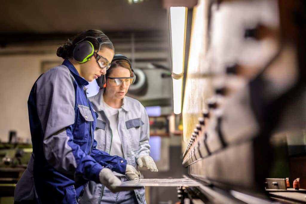 Two female apprentices wearing protective equipment work together on metal fabrication machinery, highlighting skills development and career opportunities in the defence industry.